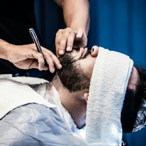An adult man receiving a precise beard trim in a barbershop with a warm towel treatment.