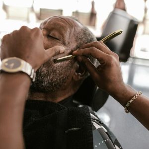 A close-up shot of a barber using a straight razor to trim a customer's beard in a barbershop.
